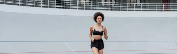 Young african american woman in black sportswear jogging on stadium, banner — Stock Photo
