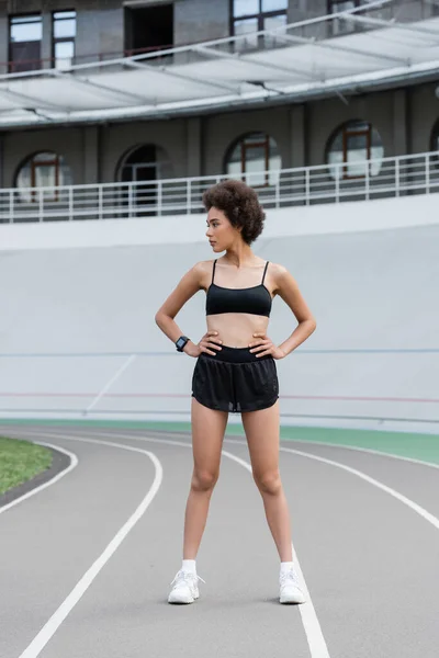 Full length of african american woman in black sportswear and white sneakers standing with hands on hips on stadium - foto de stock