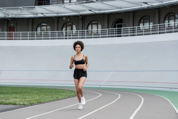 Full length of slender african american woman in black sportswear jogging on city stadium — Photo de stock