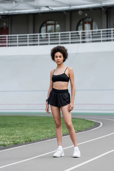 Full length of african american woman in black sports bra and shorts looking at camera on stadium - foto de stock