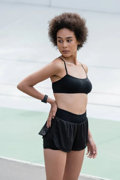 Young african american sportswoman with smart watch holding hand on hip on stadium - foto de stock