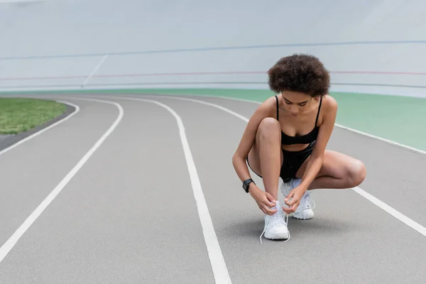 African american sportswoman tying shoelaces of sneaker on stadium — Foto stock