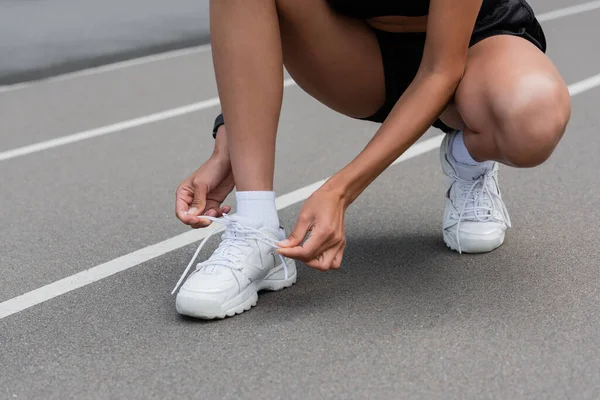 Cropped view of african american sportswoman tying shoelaces of white sneaker on stadium — Foto stock