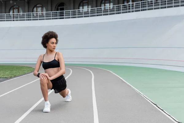 Young african american sportswoman doing lunges on running track of stadium — Stock Photo