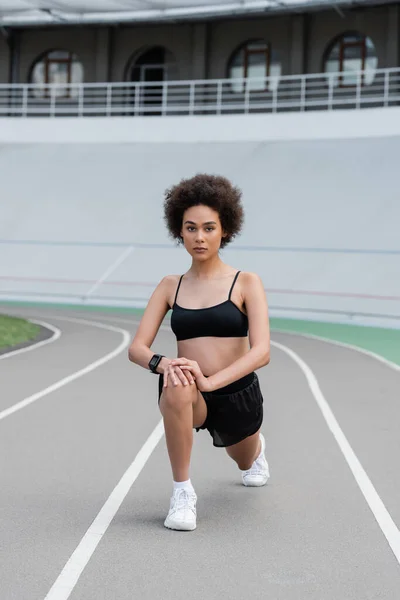 African american sportswoman doing lunges and looking at camera on stadium - foto de stock