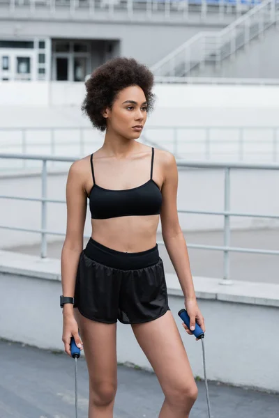 African american sportswoman in black top holding jump rope on stadium — Stockfoto