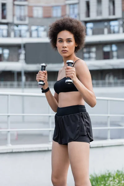 African american sportswoman in top and shorts training with dumbbells on stadium — Fotografia de Stock