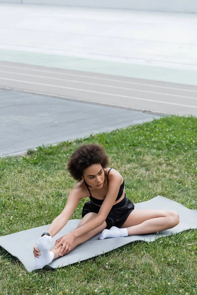 African american sportswoman stretching leg on fitness mat on stadium — Stock Photo