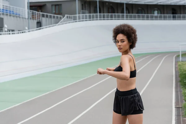 African american sportswoman in shorts and top looking at camera while stretching on stadium — Stock Photo