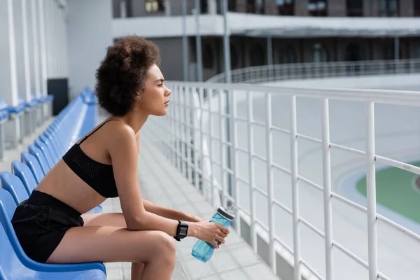 Side view of african american sportswoman holding sports bottle while sitting on stadium — Stockfoto