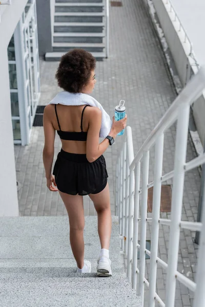 Side view of african american sportswoman with towel and sports bottle walking on stairs on stadium - foto de stock