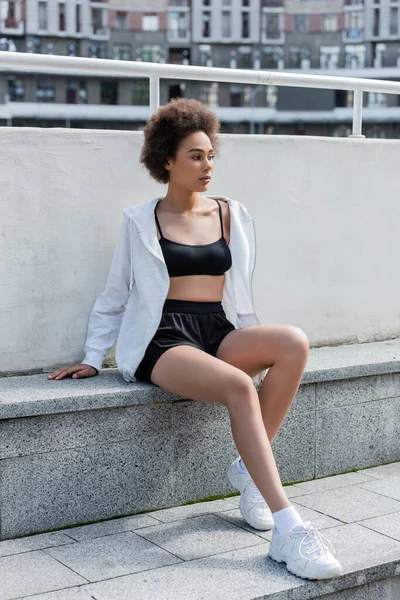 Young african american sportswoman in sports jacket and top sitting on stadium — Stockfoto