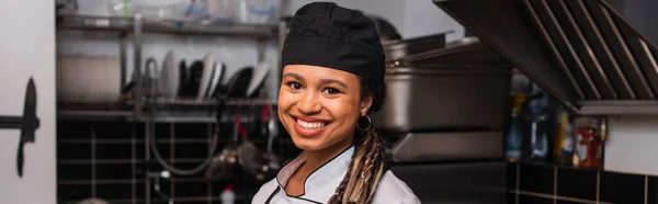 Happy and young african american chef in hat smiling in professional kitchen, banner - foto de stock