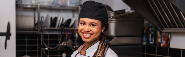 Happy and young african american chef in hat smiling in professional kitchen, banner - foto de stock
