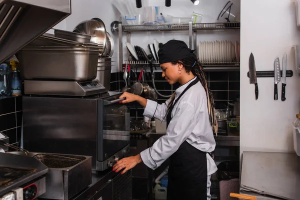 Side view of young african american chef in hat standing near convection oven in kitchen — Fotografia de Stock