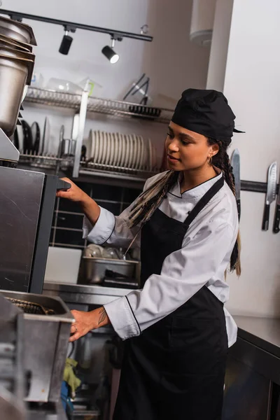 Tattooed young african american chef in hat standing near convection oven in kitchen - foto de stock