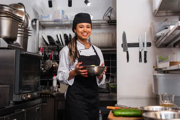 Cheerful and young african american chef holding bowl with salad in professional kitchen — Photo de stock