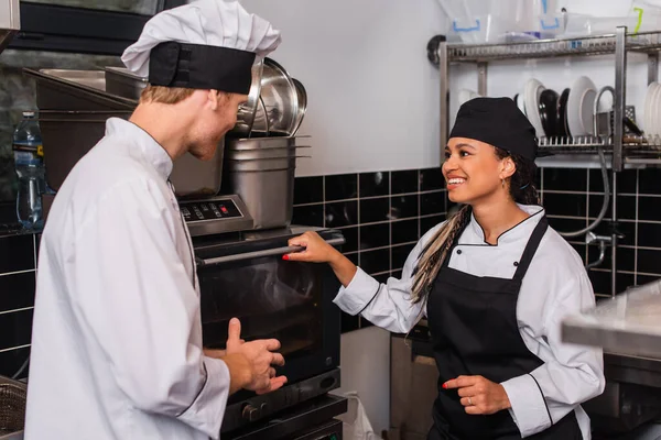 Chef in hat and uniform looking at happy african american colleague near convection oven — Foto stock