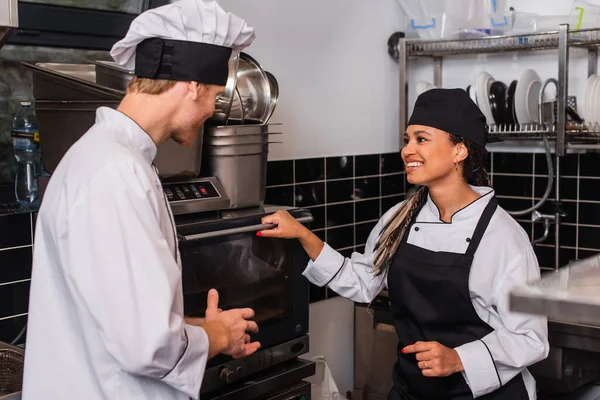 Chef in hat and uniform looking at happy african american colleague near convection oven — Stock Photo
