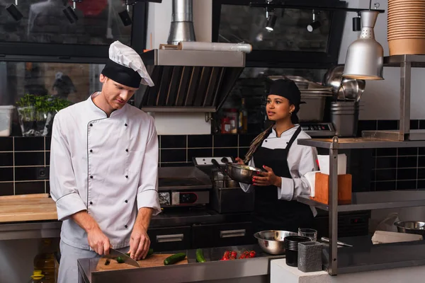 African american woman in apron holding bowl and looking at chef cutting cucumber in kitchen — стокове фото