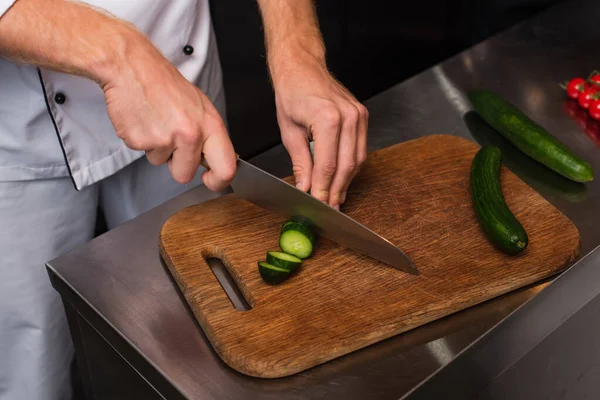 Cropped view of chef cutting cucumber on wooden cutting board - foto de stock