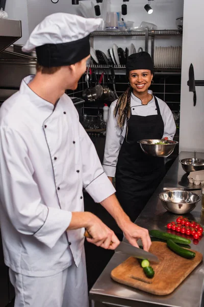 Happy african american sous chef looking at chef cutting cucumber in kitchen — стоковое фото