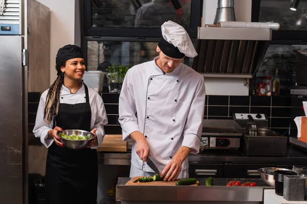 African american woman in apron holding bowl with lettuce and looking at chef preparing meal in kitchen — Stock Photo