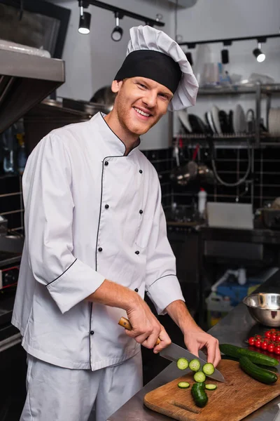 Happy young chef in uniform cutting fresh cucumber in professional kitchen — Photo de stock