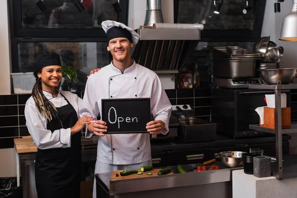 Happy young chef in uniform holding chalkboard with open lettering near african american colleague in kitchen — стоковое фото