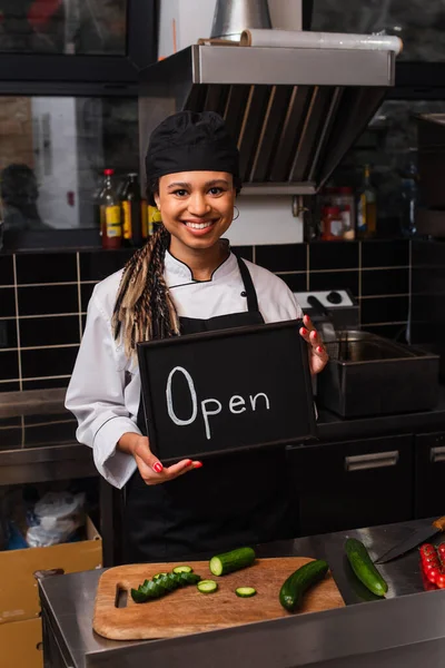 Happy african american chef holding chalkboard with open lettering in kitchen — стоковое фото