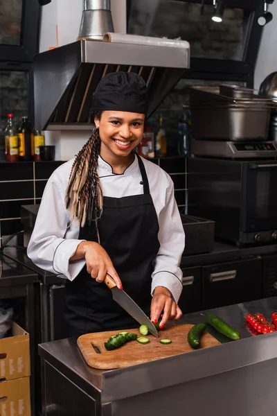 Cheerful african american woman in apron cutting cucumber in modern kitchen — стоковое фото