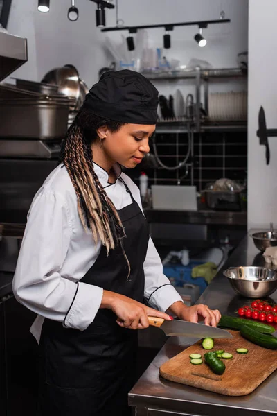 Side view of african american woman in apron cutting cucumber in modern kitchen — Stockfoto