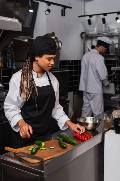 African american sous chef in apron taking cherry tomatoes while cooking in modern kitchen — Fotografia de Stock