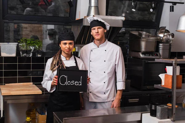 Young african american woman holding chalkboard with closed lettering near chef in kitchen — Stock Photo