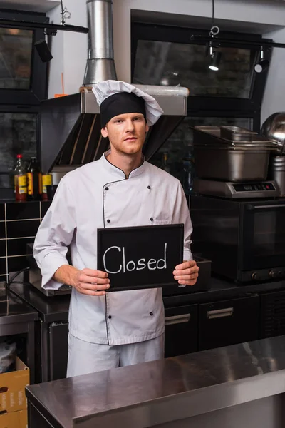Young chef in uniform holding chalkboard with closed lettering in kitchen — Photo de stock