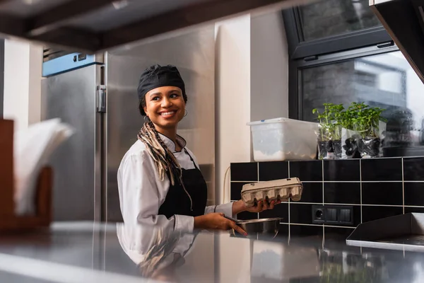 Cheerful african american chef in apron holding egg carton and bowl in kitchen — Stock Photo