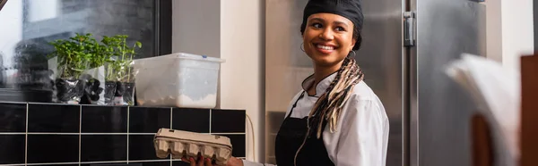 Happy african american chef in apron holding egg carton in kitchen, banner — Stock Photo