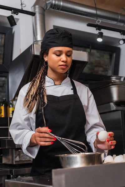 Young african american chef in apron holding egg near bowl and whisk in kitchen — Stockfoto
