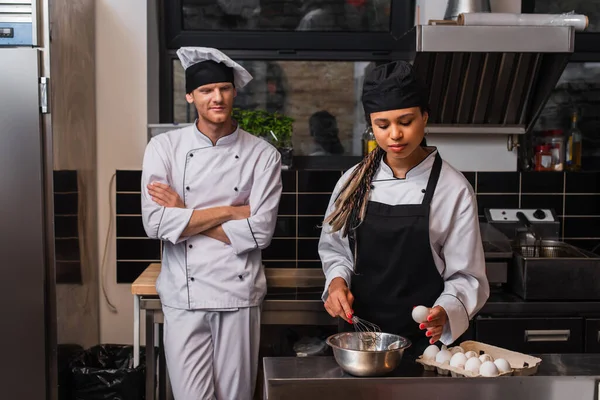 Young african american sous chef in apron holding egg near bowl and whisk while cooking near chef in kitchen — Stock Photo