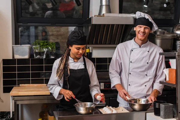 Young african american sous chef in apron holding egg near bowl and whisk while cooking near happy chef in kitchen — Stock Photo