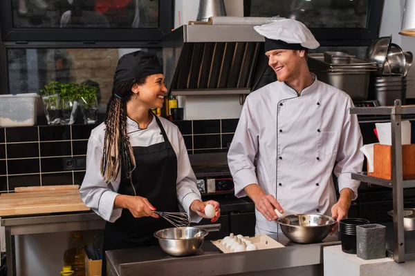 Cheerful interracial chefs holding raw eggs while cooking together in kitchen — стоковое фото