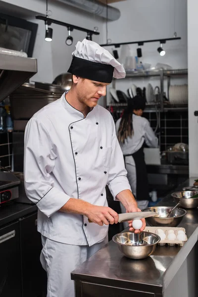 Chef in uniform holding knife near raw egg above bowl while cooking in kitchen — стоковое фото