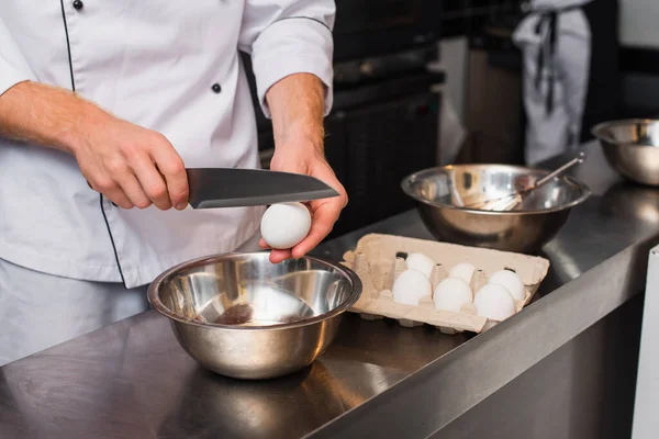 Cropped view of chef in uniform holding knife near raw egg above bowl while cooking in kitchen — Stock Photo