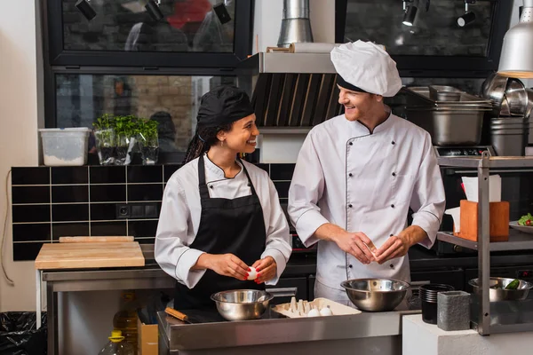 Cheerful multiethnic chefs smiling while holding raw eggs and cooking in kitchen — стоковое фото