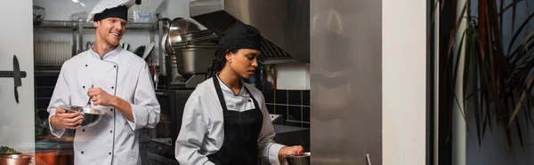 Cheerful chef whisking eggs and looking at african american colleague in kitchen, banner — стоковое фото