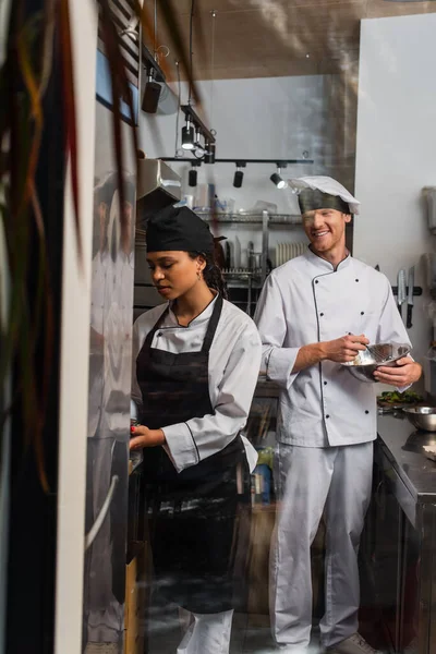 Smiling chef whisking eggs and looking at african american colleague in kitchen — стоковое фото