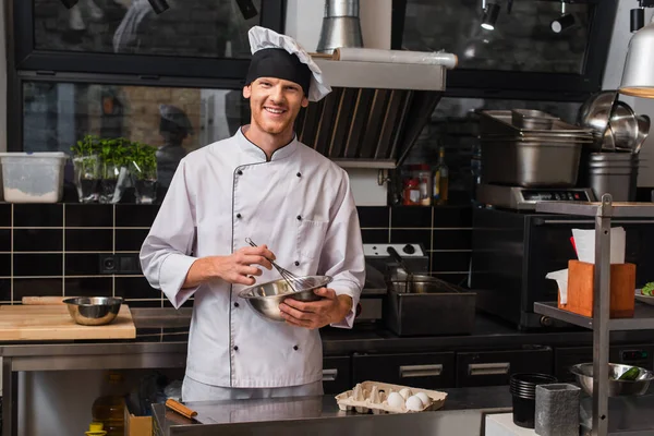 Smiling chef whisking eggs and looking at camera near egg box — Foto stock