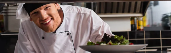 Happy chef holding plate with freshly cooked salad in professional kitchen, banner — Photo de stock