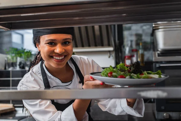 Happy african american chef holding plate with freshly cooked salad in professional kitchen — Stock Photo