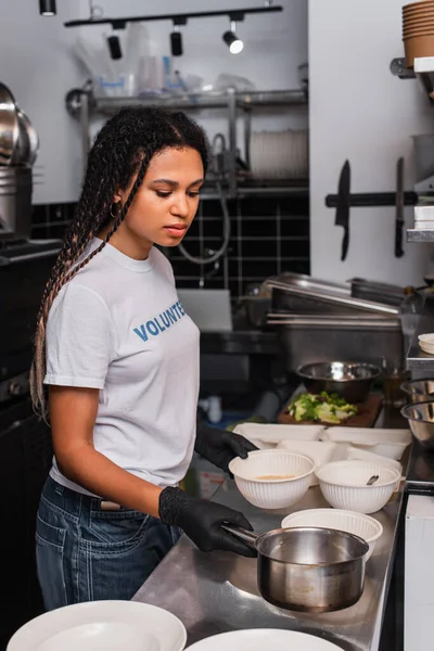African american woman in t-shirt with volunteer lettering holding saucepan near plastic bowls — Stockfoto